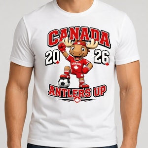May include: A white t-shirt featuring a cartoon moose mascot wearing a red soccer uniform with a maple leaf, standing with a soccer ball. The graphic includes the text CANADA, 2026, and ANTLERS UP in bold red and black lettering with maple leaf accents.