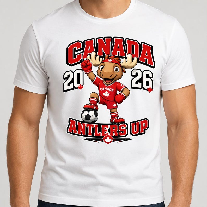 May include: A white t-shirt featuring a cartoon moose mascot wearing a red soccer uniform with a maple leaf, standing with a soccer ball. The graphic includes the text CANADA, 2026, and ANTLERS UP in bold red and black lettering with maple leaf accents.