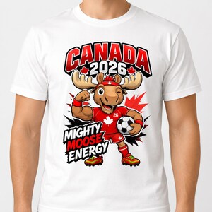 May include: A white t-shirt featuring a cartoon moose wearing a red sports uniform, a headband, and wristbands. The moose is flexing one arm and holding a soccer ball. Text reads CANADA 2026 and MIGHTY MOOSE ENERGY with red maple leaf accents.