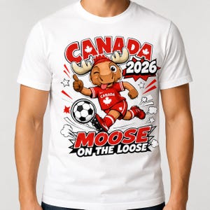 May include: A white t-shirt featuring a cartoon moose wearing a red soccer uniform with a maple leaf, kicking a soccer ball. The graphic includes the text CANADA 2026 and MOOSE ON THE LOOSE, with red accents, stars, and motion lines surrounding the character.
