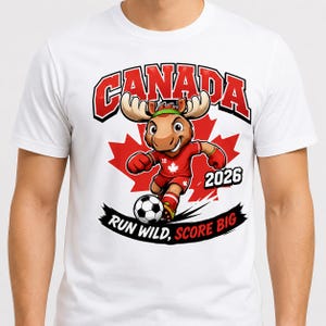 May include: A white t-shirt featuring a graphic of a cartoon moose playing soccer. The moose wears a red jersey with a maple leaf, and the design includes the text CANADA, 2026, and RUN WILD, SCORE BIG, set against a large red maple leaf background.