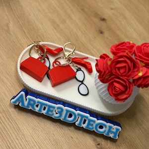 Pode incluir: Dois chaveiros vermelhos em forma de bolsa com detalhes dourados e um par de óculos em preto e branco repousam sobre uma bandeja oval branca. Um pequeno vaso branco com rosas vermelhas está à direita. O texto azul e branco diz "ARTES3DTECH".
