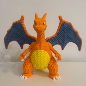 May include: A handmade orange and blue crochet Charizard figure. The figure has outstretched wings with blue and orange detailing, a yellow belly, and white claws. The figure is standing upright and is made of yarn.