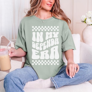 May include: Sage green t-shirt with the text "IN MY DEPENDRA ERA" in white, retro-style font. The shirt features a checkerboard pattern above and below the text. The model is wearing blue jeans.