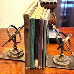 May include: A set of four vintage books held between two brass armillary sphere bookends. The leftmost book is titled Peter Pan and features a black bird illustration. The other three books have plain green, black, and maroon covers respectively.