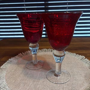 May include: Two wine glasses featuring deep red, textured glass bowls with a distinct crackled appearance. Each glass is supported by a clear, smooth glass stem and a circular base, providing a contrast between the vibrant red top and the base.