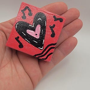 Puede incluir: Un pequeño lienzo cuadrado rojo con un diseño de corazón negro y notas musicales. El corazón tiene un corazón interior rosa y detalles de costura blancos. Líneas onduladas negras en la parte inferior.