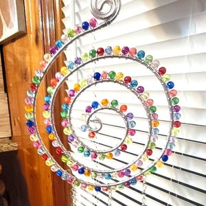 May include: A spiral-shaped hanging ornament constructed from silver-toned wire, adorned with numerous multi-colored translucent beads. The structure features a central coil design with three dangling chains at the bottom, each ending in a clear bead.