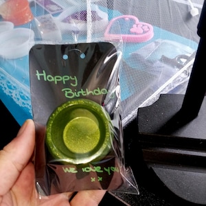 Op de afbeelding: Een groene, glinsterende hars ringhouder in een doorzichtige plastic verpakking. De kaart leest "Happy Birthday" en "We love you" in groene stift. De houder is rond met een ondiepe schaal.