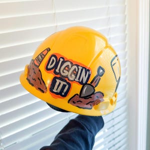 May include: A bright yellow hard hat featuring a hand-painted design of brown dirt piles, an orange traffic cone, and a small shovel. The text "DIGGIN IT!" is written in bold, dark letters with a light outline centered on the front of the helmet.