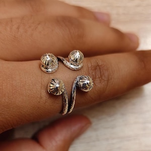 Könnte beinhalten: Ein silberfarbener Ring mit einem einzigartigen Design. Der Ring hat vier dekorative, kugelförmige Elemente mit aufwendigen Details. Der Ring umschlingt den Finger und schafft ein unverwechselbares Accessoire. Der Ring wird auf einer Hand gezeigt.