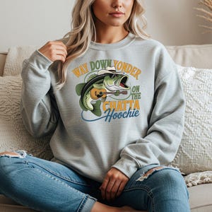 May include: A light grey crewneck sweatshirt featuring a graphic of a green bass fish wearing a white cowboy hat and sunglasses while playing a brown acoustic guitar. The text reads Way Down Yonder On The Chatta Hoochie in green and yellow.
