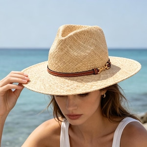 May include: A light beige straw fedora hat featuring a structured crown and a wide brim. The hat is accented with a thin, dark brown leather band that wraps around the base of the crown, finished with a small, polished gold-tone metal horsebit detail.