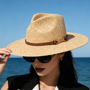 May include: A light tan woven straw fedora hat featuring a thin brown leather band with a gold-toned metal buckle detail. The hat has a structured crown and a wide brim, providing a classic look suitable for sunny weather and outdoor settings.