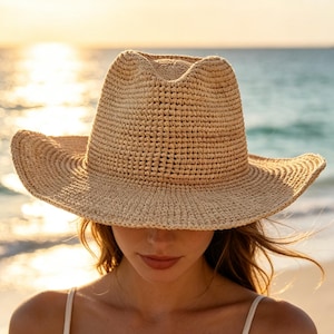 May include: A beige straw fedora hat with a structured crown and a wide, slightly upturned brim. The hat features a tightly woven texture throughout, providing a classic look. It is paired with a simple white sleeveless top with thin straps.