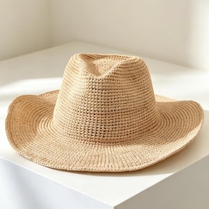 May include: A light beige fedora hat crafted from a woven straw-like material. The hat features a pinched crown with a central crease and a wide, slightly upturned brim. The texture is consistent throughout, showing a dense, uniform crochet pattern.