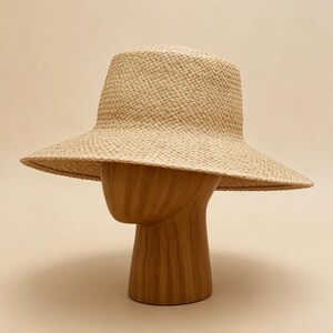 May include: A light beige woven straw hat with a structured crown and a wide, slightly downturned brim. The hat features a consistent, tight basket-weave pattern throughout, showcasing a natural, neutral colour palette and a clean, simple silhouette.