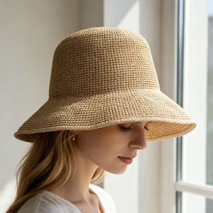 May include: A beige bucket hat crafted from a tightly woven, textured material. The hat features a rounded crown and a wide, downward-sloping brim that provides coverage. The construction shows a consistent, uniform stitch pattern throughout the design.