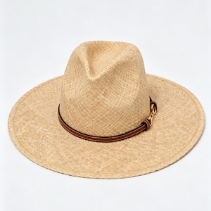 May include: A light beige woven straw fedora hat with a wide brim and a pinched crown. The hat features a thin brown leather-like band encircling the base of the crown, accented with a small gold-toned metal chain link detail on the side.