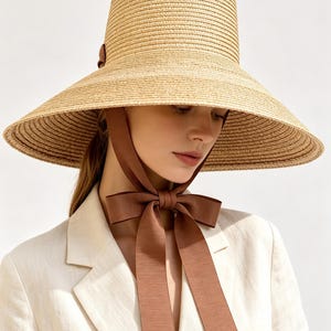 May include: A wide-brimmed straw hat in a natural tan colour featuring a tall, structured crown and a brown ribbon chin strap that ties into a bow under the chin. The hat is paired with a light beige blazer with a notched lapel collar.