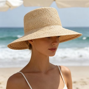 May include: A light beige straw hat with a structured crown and a wide, slightly downturned brim. The hat features a woven texture throughout, providing a natural tan colour. It is paired with a simple white sleeveless top with thin shoulder straps.