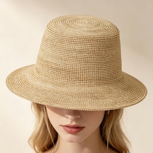 May include: A light tan straw bucket hat with a flat top and a circular woven texture. The hat features a structured crown and a downward-sloping brim that provides coverage. The material is a natural, woven fibre with a consistent, tight weave.