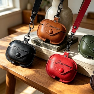 May include: A collection of leather AirPod cases in various colors, including black, brown, red, and green. Each case has a strap and a snap closure. The cases are personalized with names like "William", "Cameryn", and "Journey".