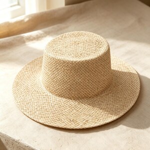 May include: A light beige straw hat featuring a flat crown and a wide, circular brim. The hat is constructed with a woven pattern throughout, showcasing a natural tan colour and a structured, cylindrical shape with a flat top surface.