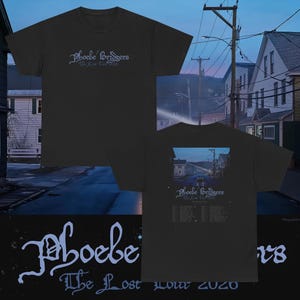 Puede incluir: Una camiseta negra de manga corta con el texto Phoebe Bridgers The Lost Tour 2020 en fuente gótica azul claro en el frente. La parte posterior muestra una escena de calle suburbana al atardecer con el mismo texto y una lista de fechas de gira.