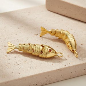 May include: Two gold-toned articulated fish pendants featuring a textured, scale-like body design. Each fish has a smooth head with a small circular orange stone set as an eye and a metal loop at the mouth for attachment to a chain or cord.