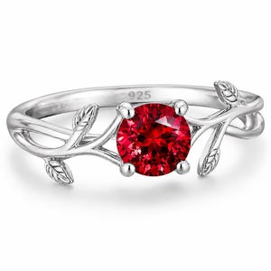 May include: A silver ring featuring a round, vibrant red gemstone set in the centre. The band is designed to resemble a delicate vine with small, etched leaves extending along both sides. The inner band is clearly stamped with the number 925.