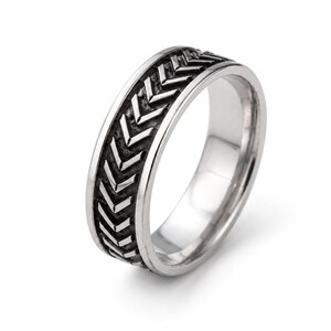 May include: A silver-toned metal ring featuring a continuous band of raised, chevron-shaped arrow patterns. The recessed areas between the arrows are darkened to provide contrast, while the inner surface of the ring is smooth and polished.