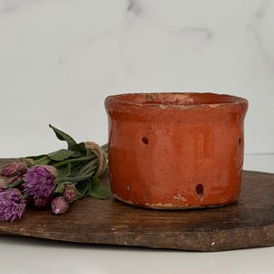 May include: A cylindrical terracotta pot with a rustic orange glaze and small drainage holes along the base, accompanied by a small bundle of purple chive blossoms tied with twine, resting on a dark, weathered wooden board.