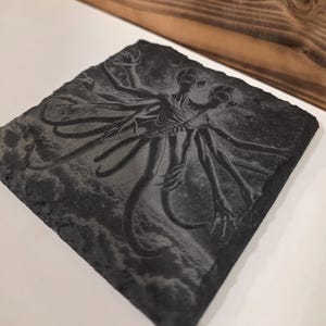 May include: A dark gray square slate coaster featuring an etched design of a two-headed alien creature with multiple arms and tentacles. The artwork is detailed and set against a cloudy background.