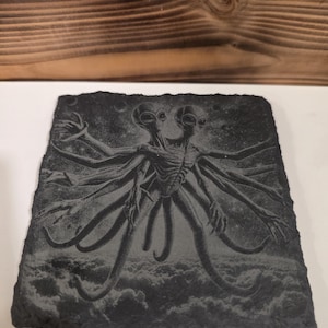 May include: A dark gray slate coaster featuring an etched image of a two-headed alien with multiple arms and a cloud-filled background. The artwork is detailed and the coaster has a rough, natural edge.