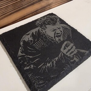 Puede incluir: Posavasos cuadrado de pizarra negra con una imagen grabada de un cantante sosteniendo un micrófono y cantando. La imagen es detallada, mostrando la expresión facial y la ropa del cantante. El posavasos está sobre una superficie blanca.