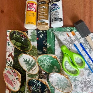 May include: A craft kit featuring five decoupage oyster shells with holiday patterns, three bottles of paint and glue, green scissors, two paintbrushes, and a foam brush. The bottles include Mod Podge Matte-Mat, DecoArt Glorious Gold, and Americana White.