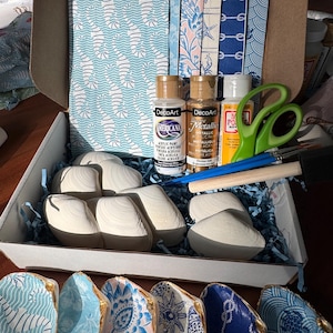 May include: A craft kit in an open cardboard box, featuring white seashells, paint, brushes, and scissors. The kit includes various patterned papers and DecoArt acrylic paints. Painted seashells with gold trim are displayed at the bottom.