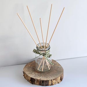 May include: A clear glass bottle containing five thin wooden reed diffuser sticks. The neck of the bottle is tied with a green and white gingham ribbon. The bottle rests on a circular, natural wood slice with visible bark edges.