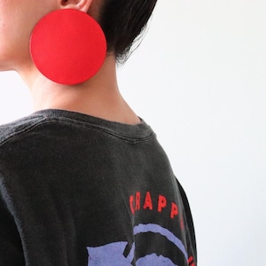 May include: A large, circular, bright red earring with a smooth, matte finish. The earring is worn on the earlobe and features a simple, flat, disc-shaped design that covers a significant portion of the side of the head, creating a bold visual statement.