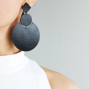 May include: A black earring featuring a diamond-shaped top piece connected to a medium-sized circle, which is attached to a larger circular base. The earring has a textured surface and is constructed from three distinct geometric shapes linked together.