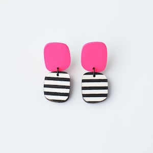 May include: A pair of pink and black striped dangle earrings. The earrings are made of wood and have a square shape.