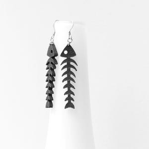 May include: A pair of dangling earrings featuring black, flat, fish skeleton silhouettes. Each earring is suspended from a silver-toned metal hook and displays a detailed, segmented spine design with a circular cutout representing the eye.