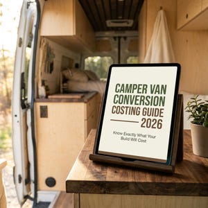 May include: A tablet showing "Camper Van Conversion Costing Guide 2026" in green text, on a wooden stand. The tablet is on a wooden surface inside a camper van, with a small potted plant visible.