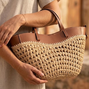 Pode incluir: Uma bolsa tote de palha tecida com alças e acabamentos em couro marrom. A bolsa tem um design de tecido aberto e texturizado e é segurada por uma pessoa vestindo uma camisa de linho de cor clara. A guarnição de couro é um marrom rico e quente.