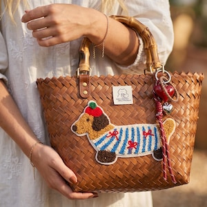 Pode incluir: Uma sacola tote marrom tecida com alças de bambu e um patch decorativo de cachorro. O patch do cachorro apresenta um dachshund vestindo uma camisa listrada com laços vermelhos e um chapéu. Um pingente de coração vermelho pende da alça.