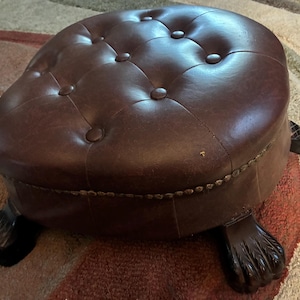 May include: A round, dark brown ottoman featuring a tufted top with button accents. The base is supported by four dark wooden feet carved into a claw-and-ball design. A row of small, metallic nailhead trim encircles the side of the seat cushion.