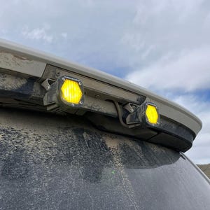 May include: Two square LED auxiliary lights with bright yellow lenses mounted to the top edge of a vehicle's rear window frame. The lights feature a metallic housing and are positioned horizontally, showing visible dust on the vehicle surface.