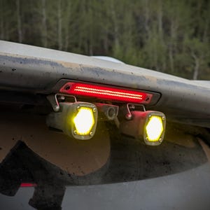 May include: Two square auxiliary lights with bright yellow hexagonal LED beams mounted beneath a vehicle's rear spoiler, positioned on either side of a central red horizontal brake light strip, featuring metallic brackets and exposed hardware.