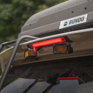 May include: A vehicle rear light assembly featuring a horizontal red LED strip light mounted above two smaller rectangular auxiliary lights with amber lenses. The assembly is attached to a beige vehicle body below a black roof box labeled BUNDU.